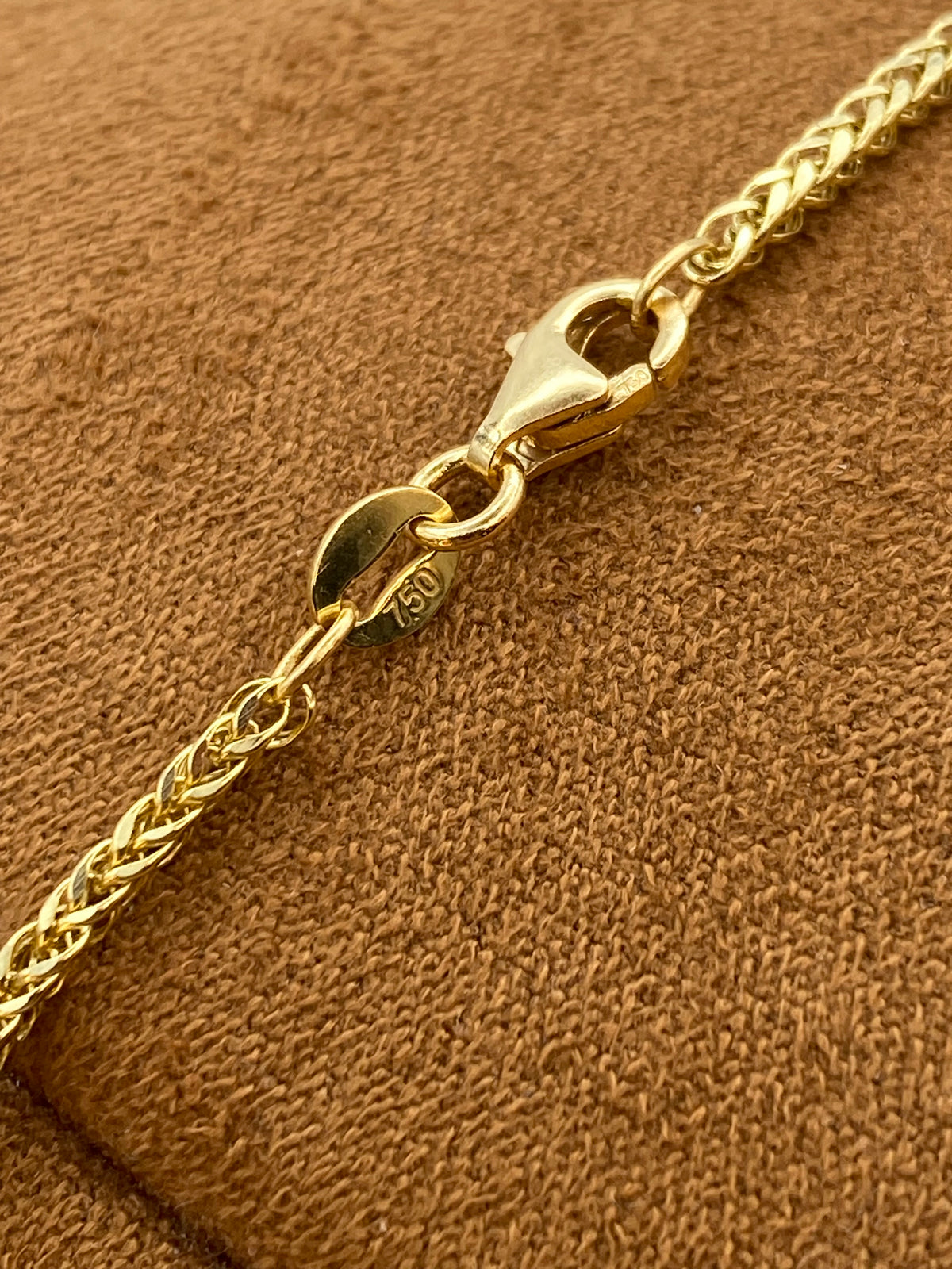 Close-up of a gold chain bracelet with a lobster clasp and a round 14k gold hallmark tag, resting on a textured brown suede surface.