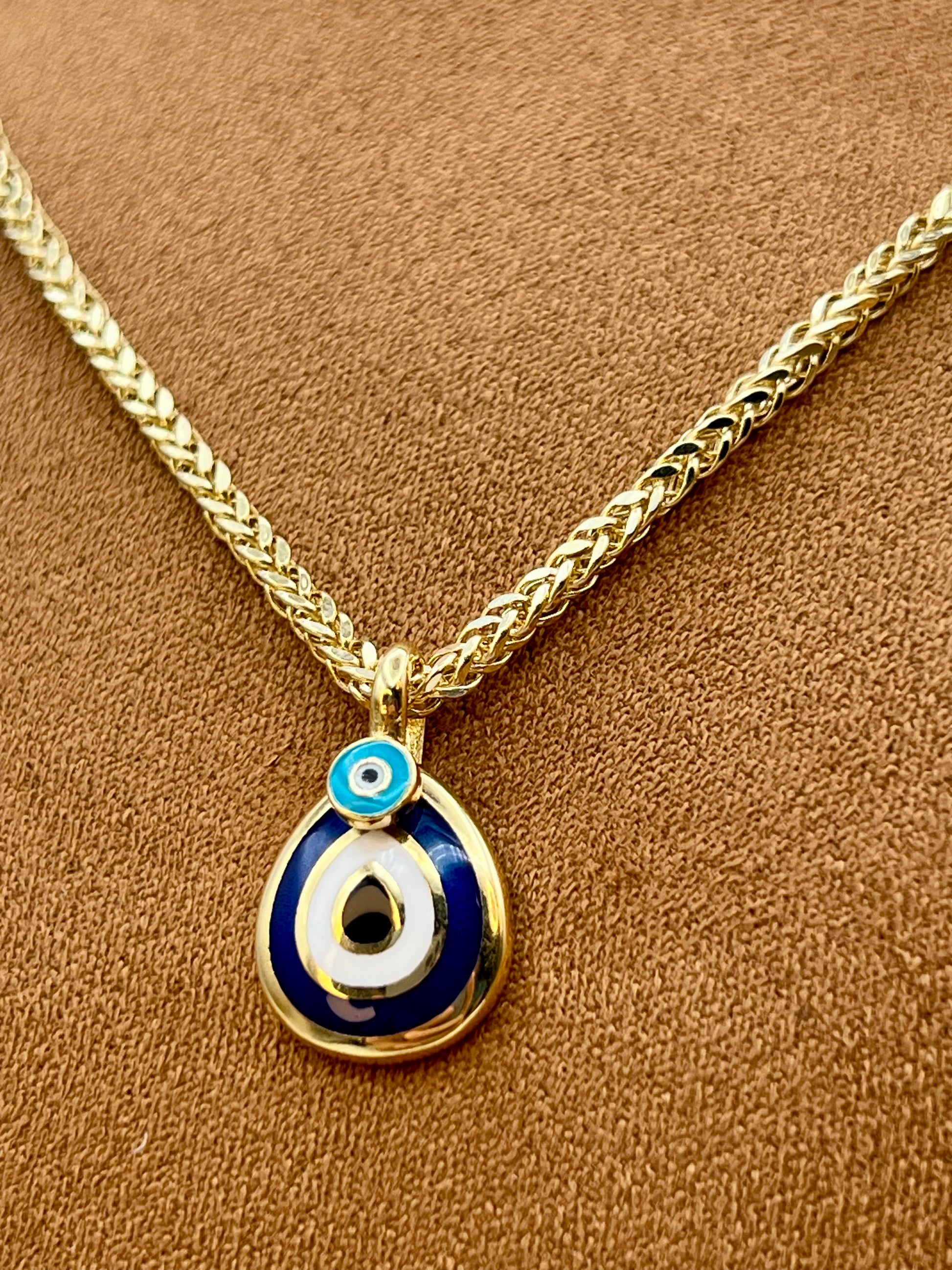 Gold-tone chain necklace with a teardrop-shaped evil eye pendant on a textured brown background.