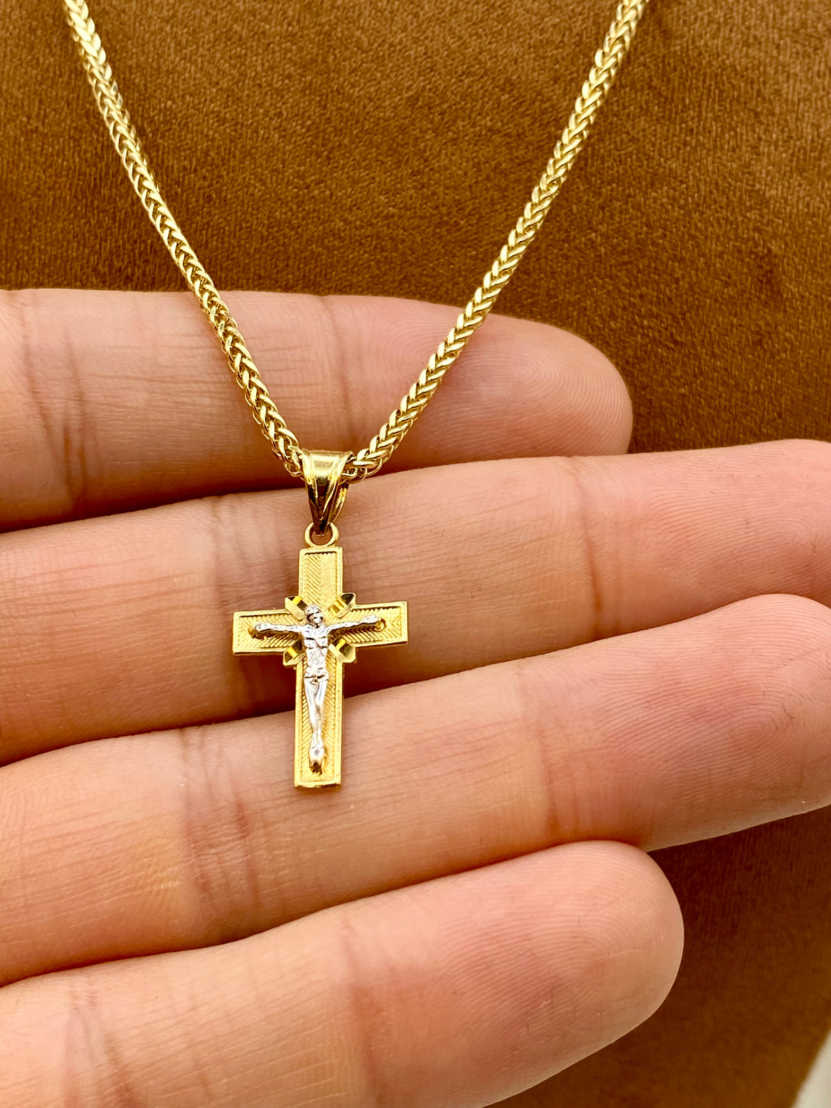 A gold cross pendant necklace with a small clear stone in the center, displayed on a person's hand against a brown textured background.