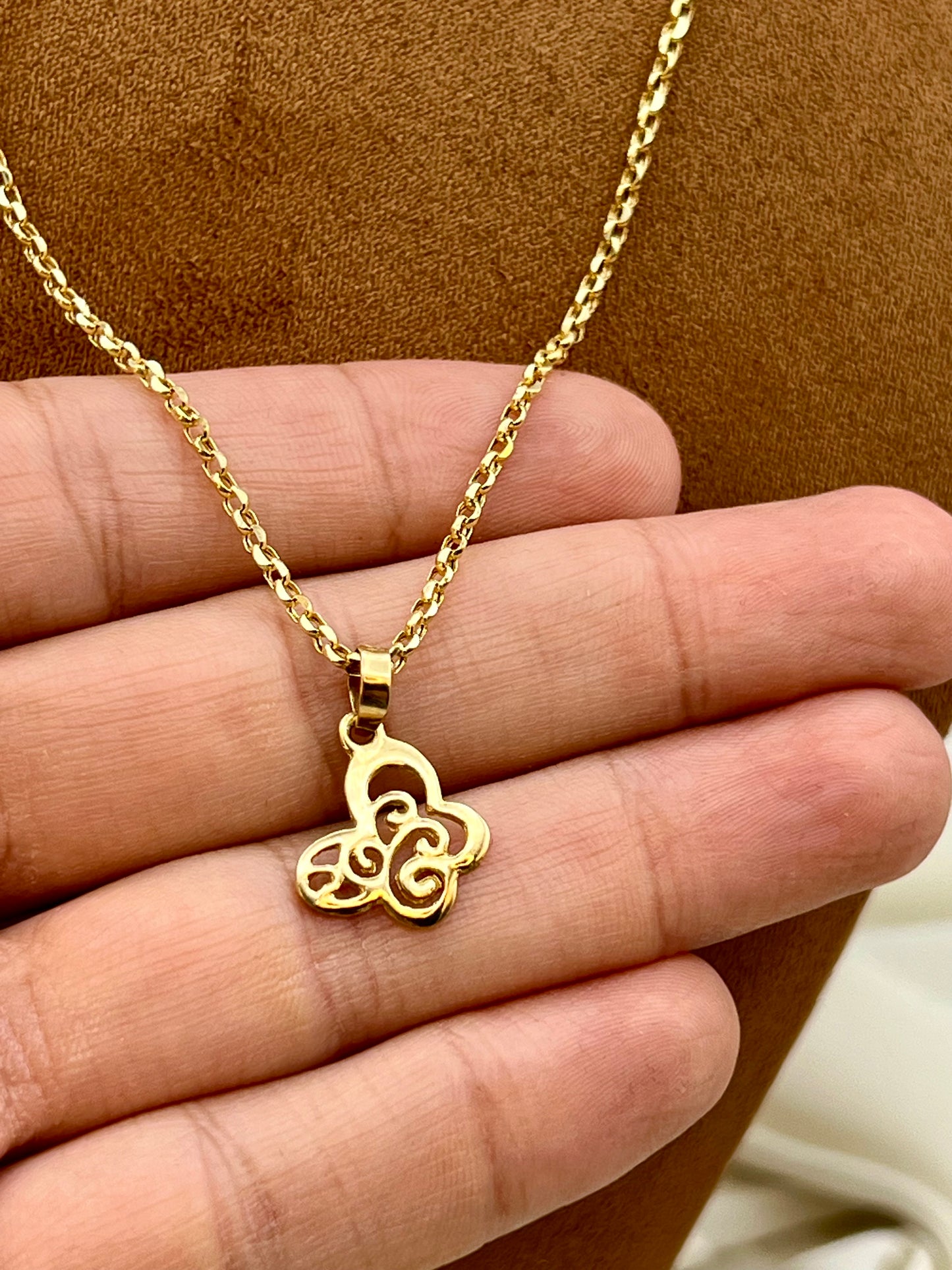 A gold necklace with a Butterfly-shaped pendant is held in a person's hand against a brown textured background.