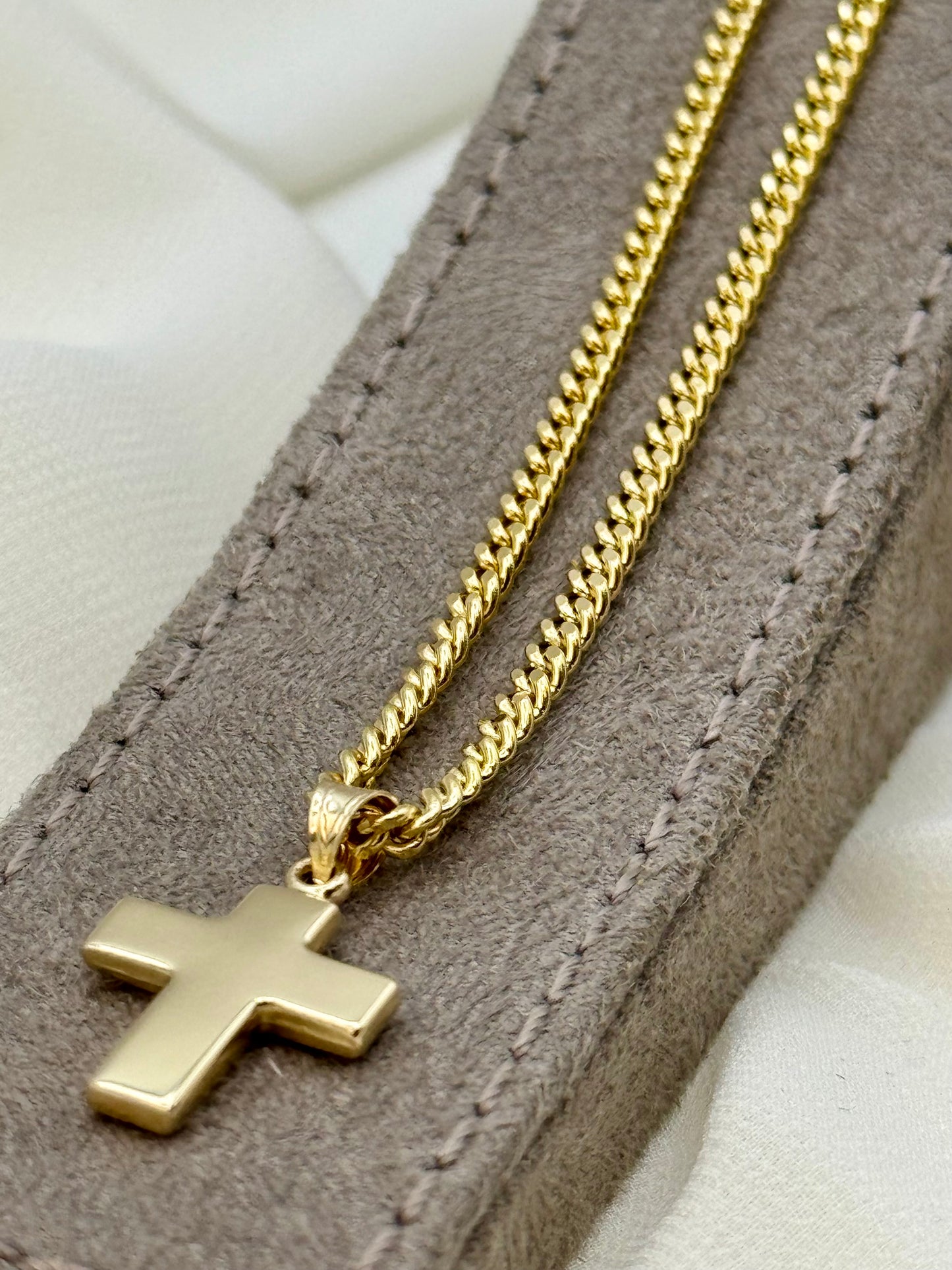 A gold cross pendant necklace on a gold curb chain, displayed on a brown suede jewelry pouch over a white fabric background.