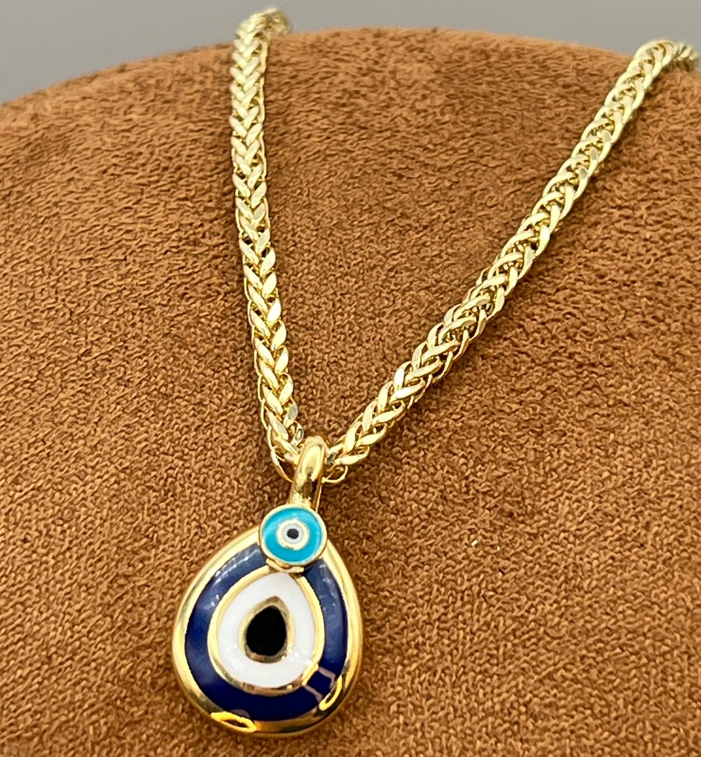 A gold chain necklace with an evil eye pendant, displayed on a brown textured jewelry stand. The pendant features blue, white, and black enamel detailing with a gold border.
