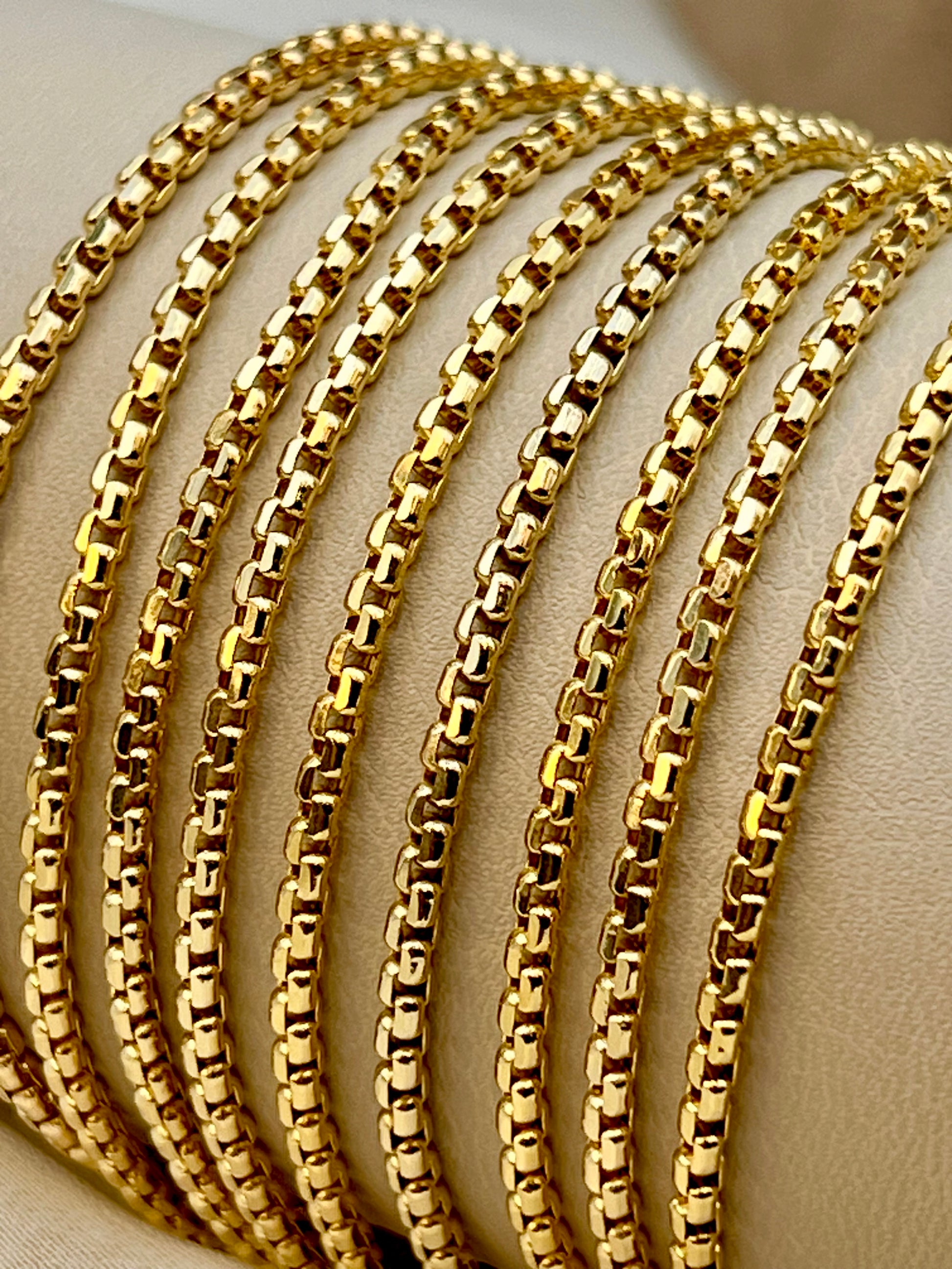 A close-up of several overlapping 18k gold-tone Box chains displayed on a beige jewelry stand.