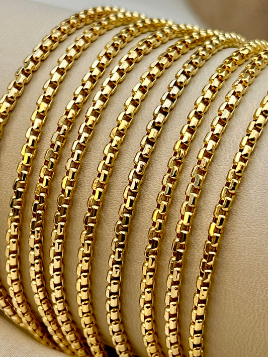 A close-up of several overlapping 18k gold-tone Box chains displayed on a beige jewelry stand.