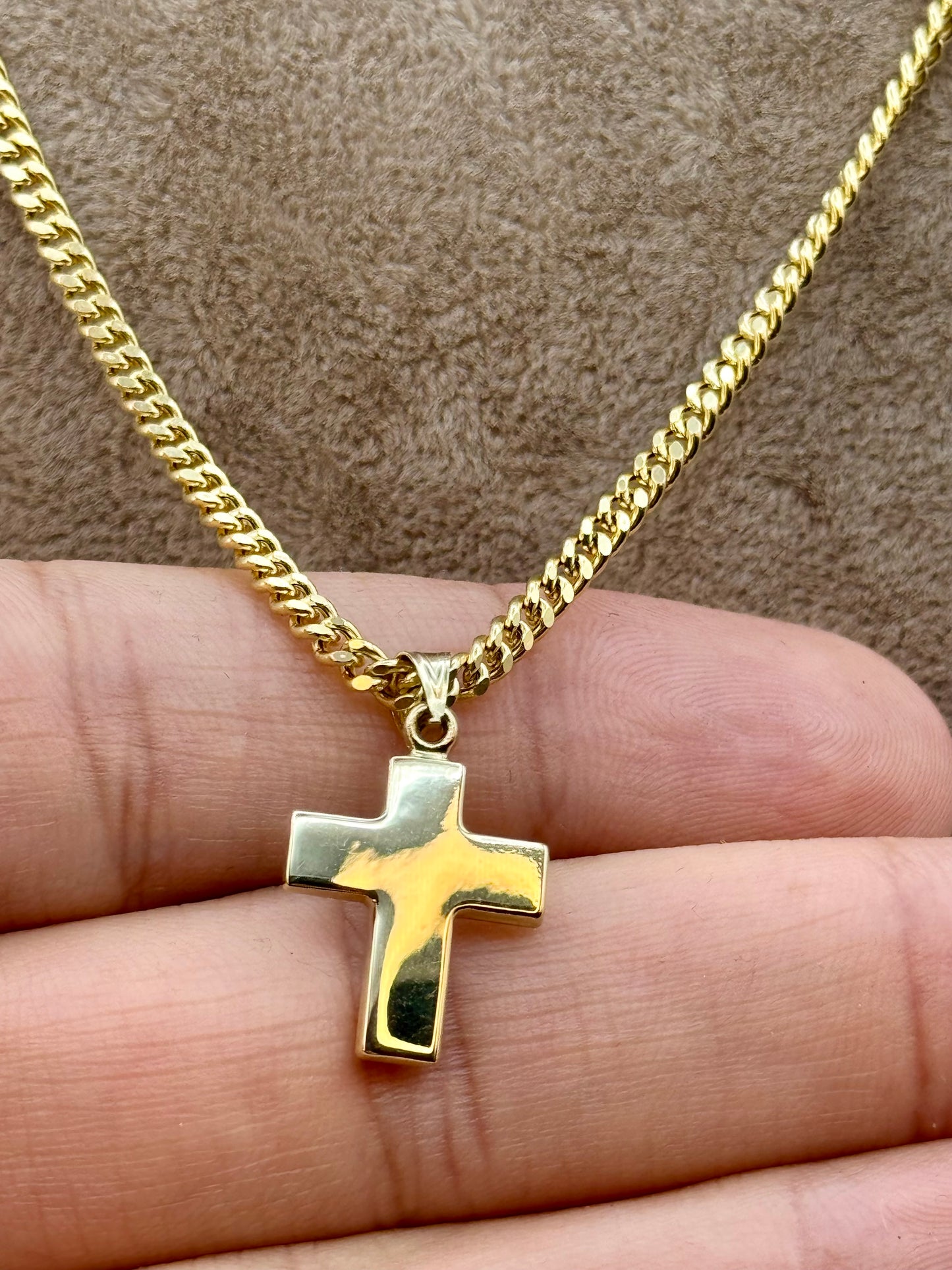 A gold cross pendant necklace on a gold chain, held in a person's hand against a brown textured background.
