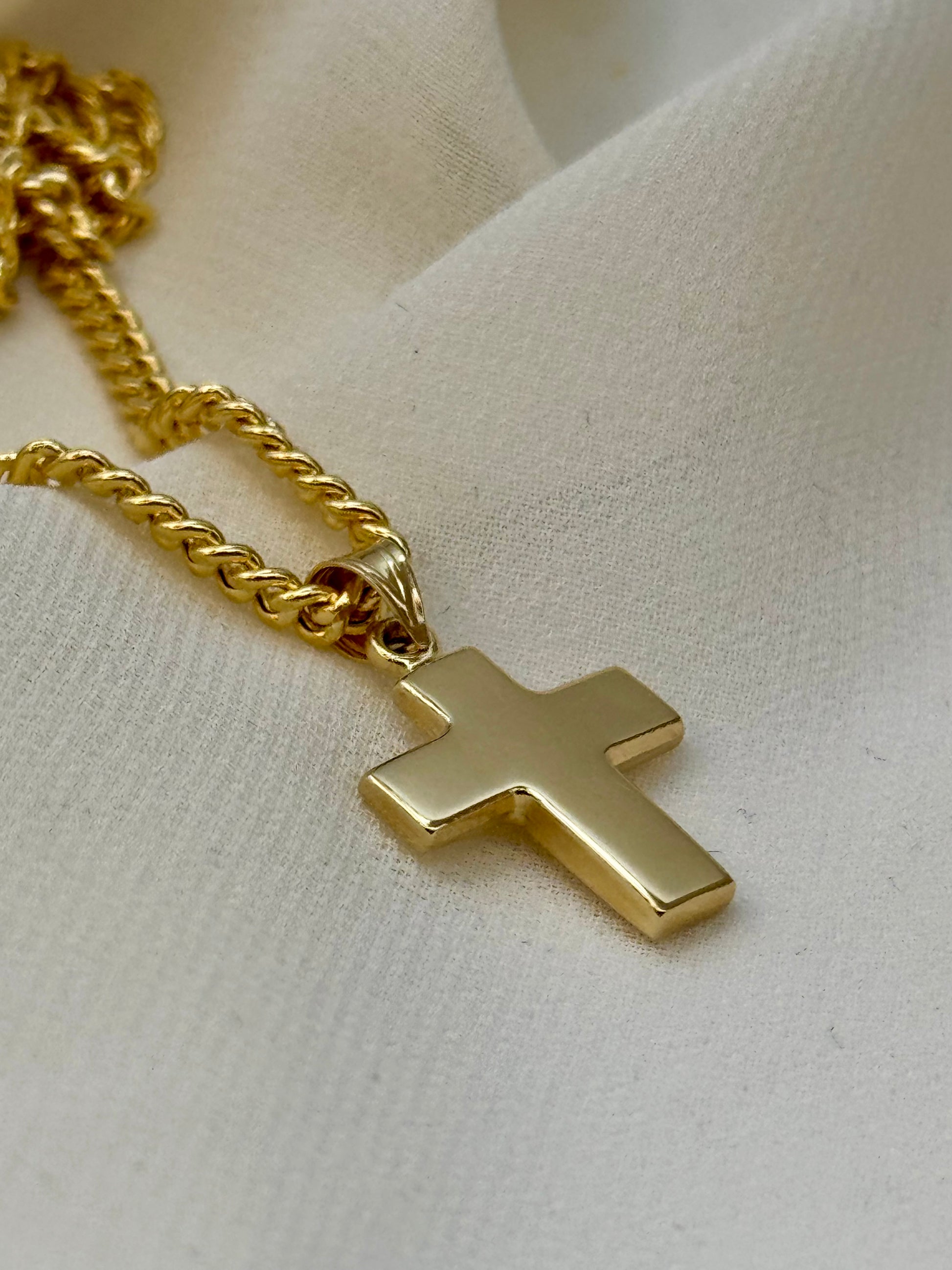 A polished gold cross pendant necklace on a gold chain, displayed on a textured off-white fabric background.