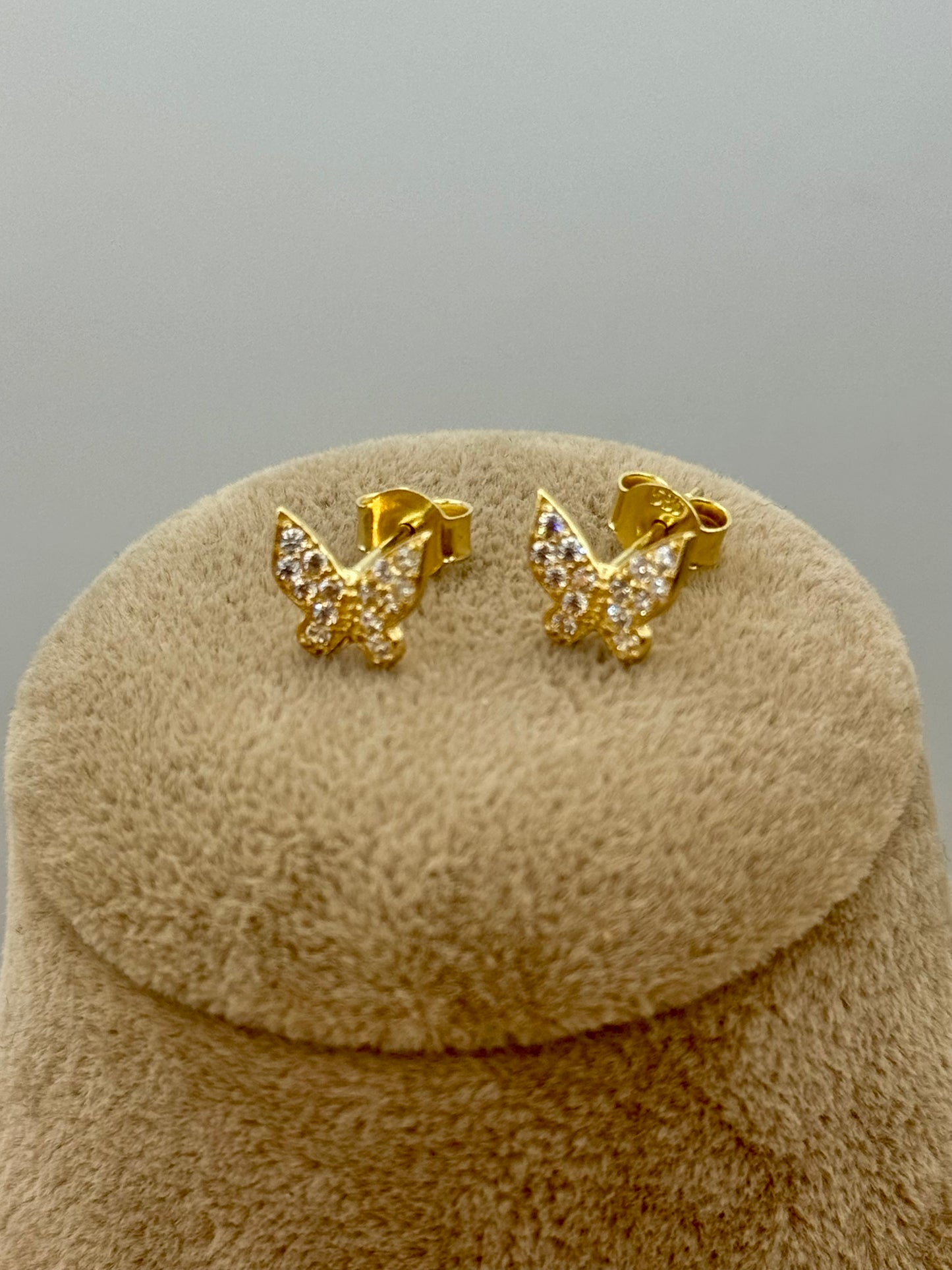A pair of gold butterfly stud earrings with clear pavé-set stones, displayed on a round beige velvet jewelry stand against a neutral gray background.