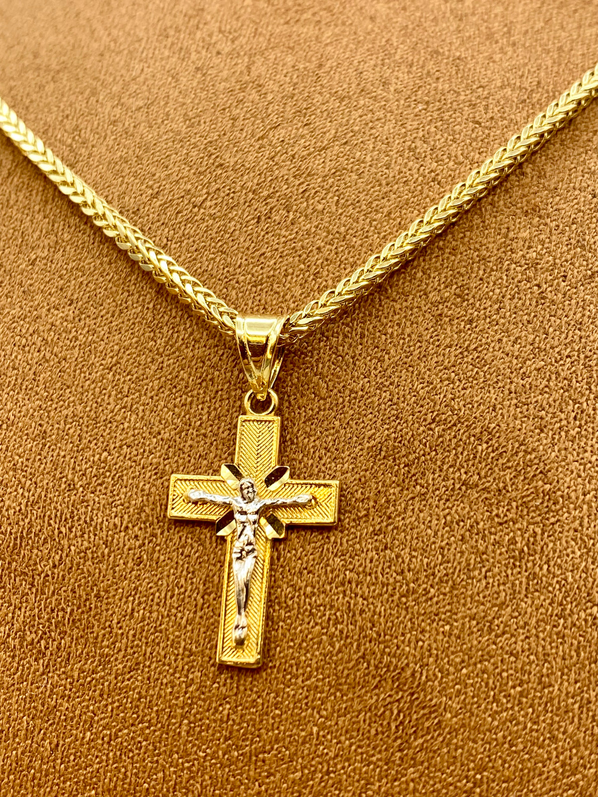 A gold crucifix pendant necklace with a detailed figure of Jesus on a textured cross, displayed on a braided gold chain against a brown textured background.