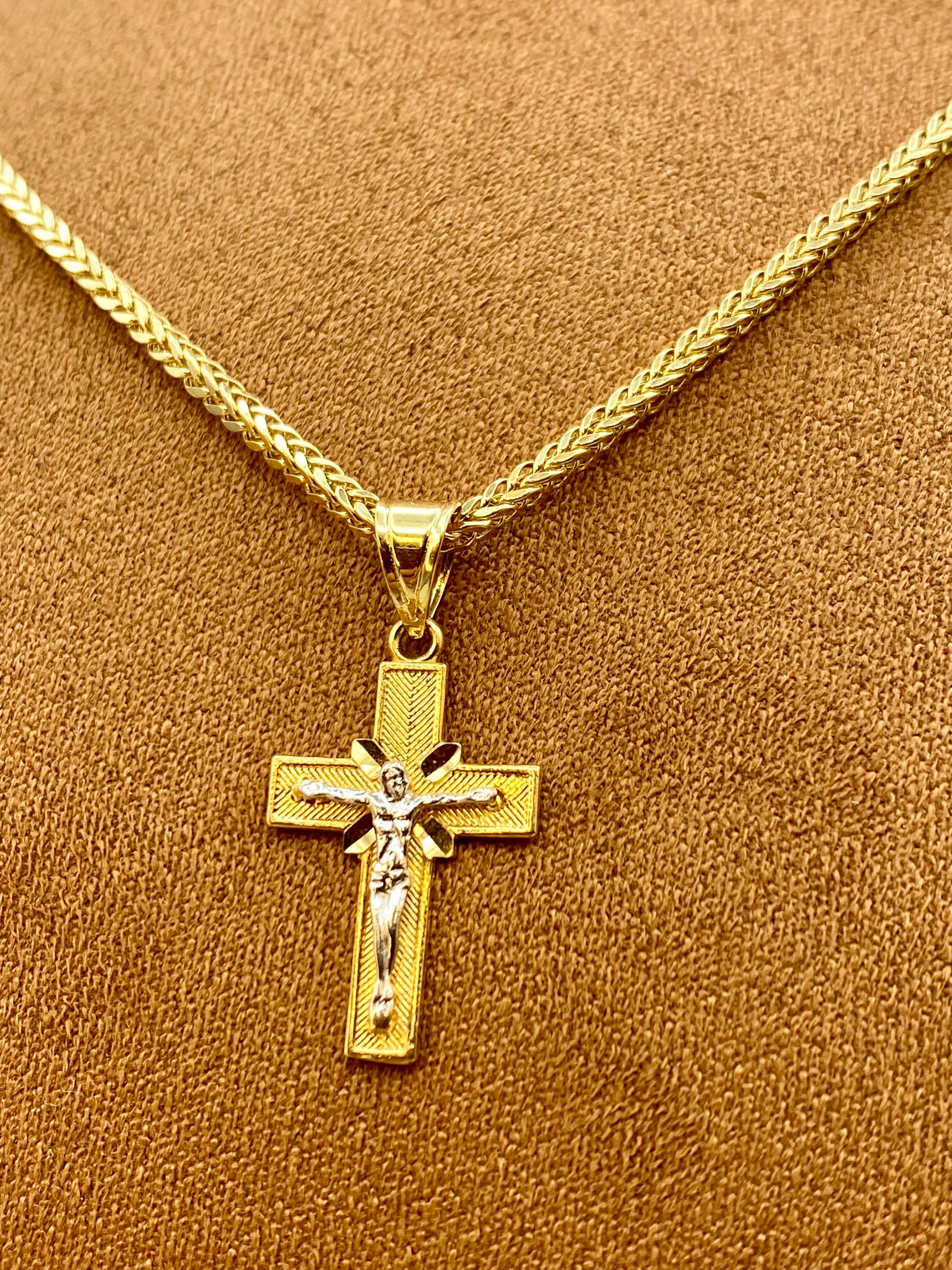 A gold crucifix pendant necklace with a detailed figure of Jesus on a textured cross, displayed on a braided gold chain against a brown textured background.