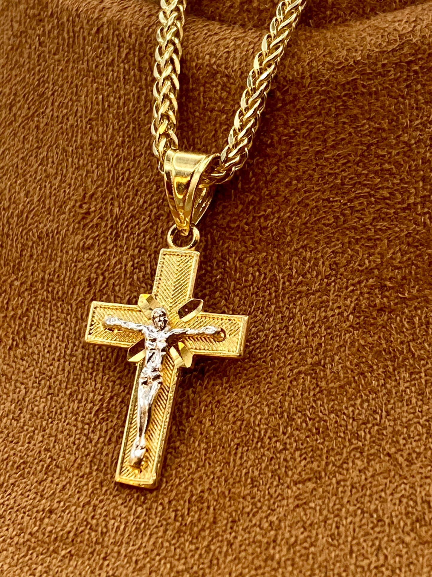 A gold cross pendant necklace with a detailed crucifix design, resting on a textured brown fabric background.