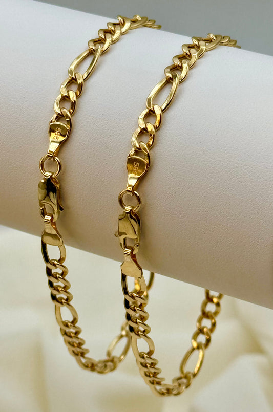 Two 14k gold figaro chain bracelets on a white arm against a beige background