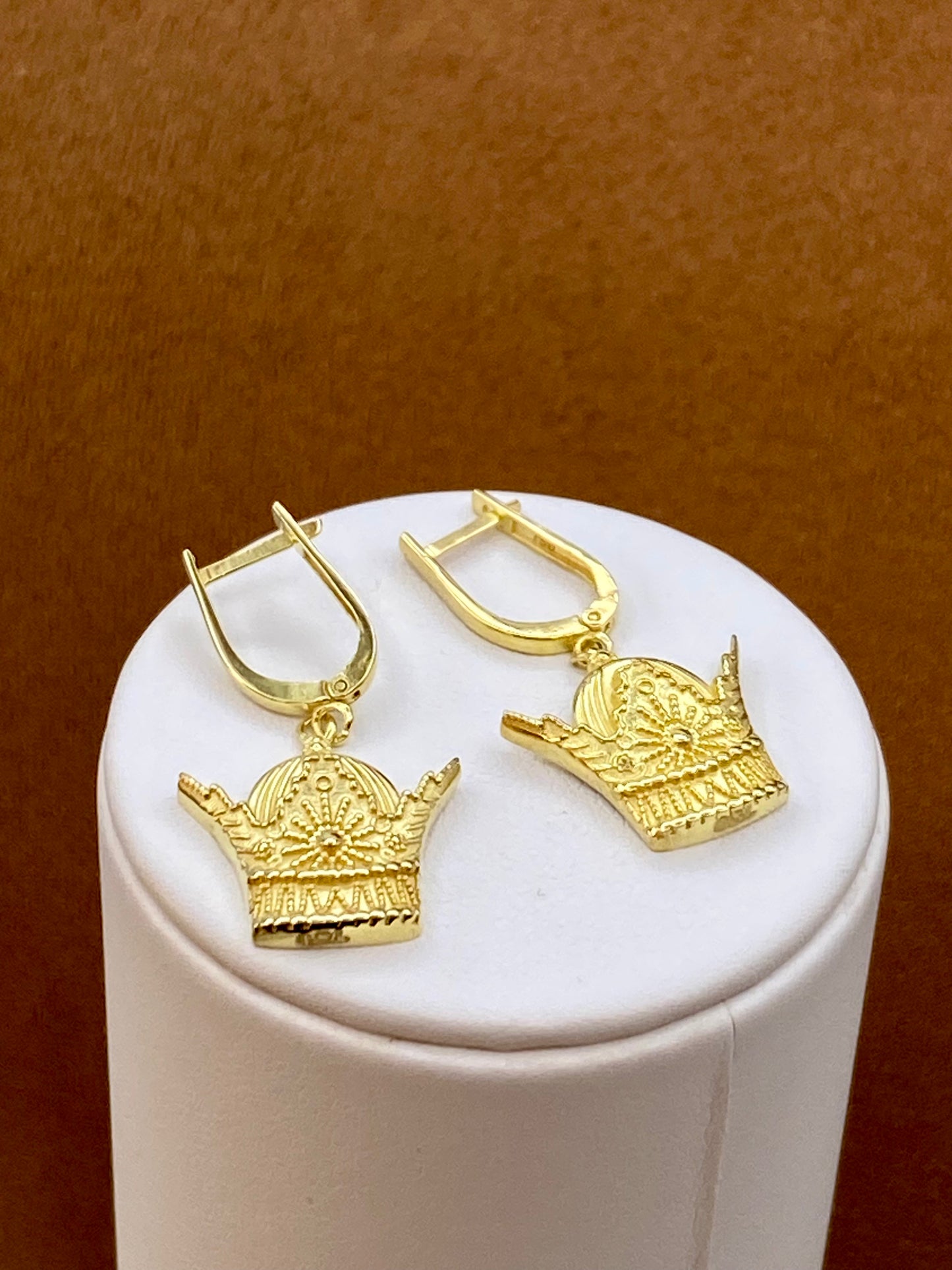 18k Gold crown-shaped earrings on a white cylindrical stand with a brown background