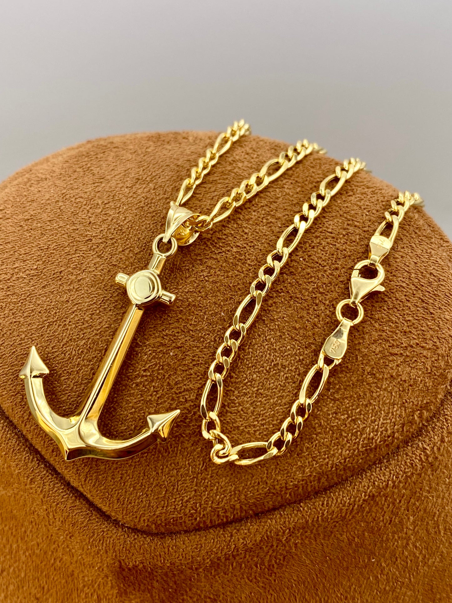 18k Gold anchor necklace and chain on a brown cushion with a gray background