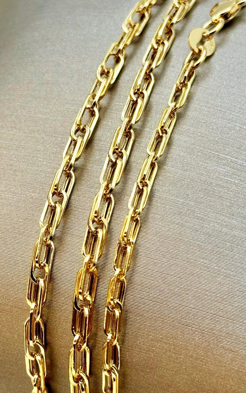 A close-up of a gold paperclip chain necklace draped diagonally on a textured beige fabric background.