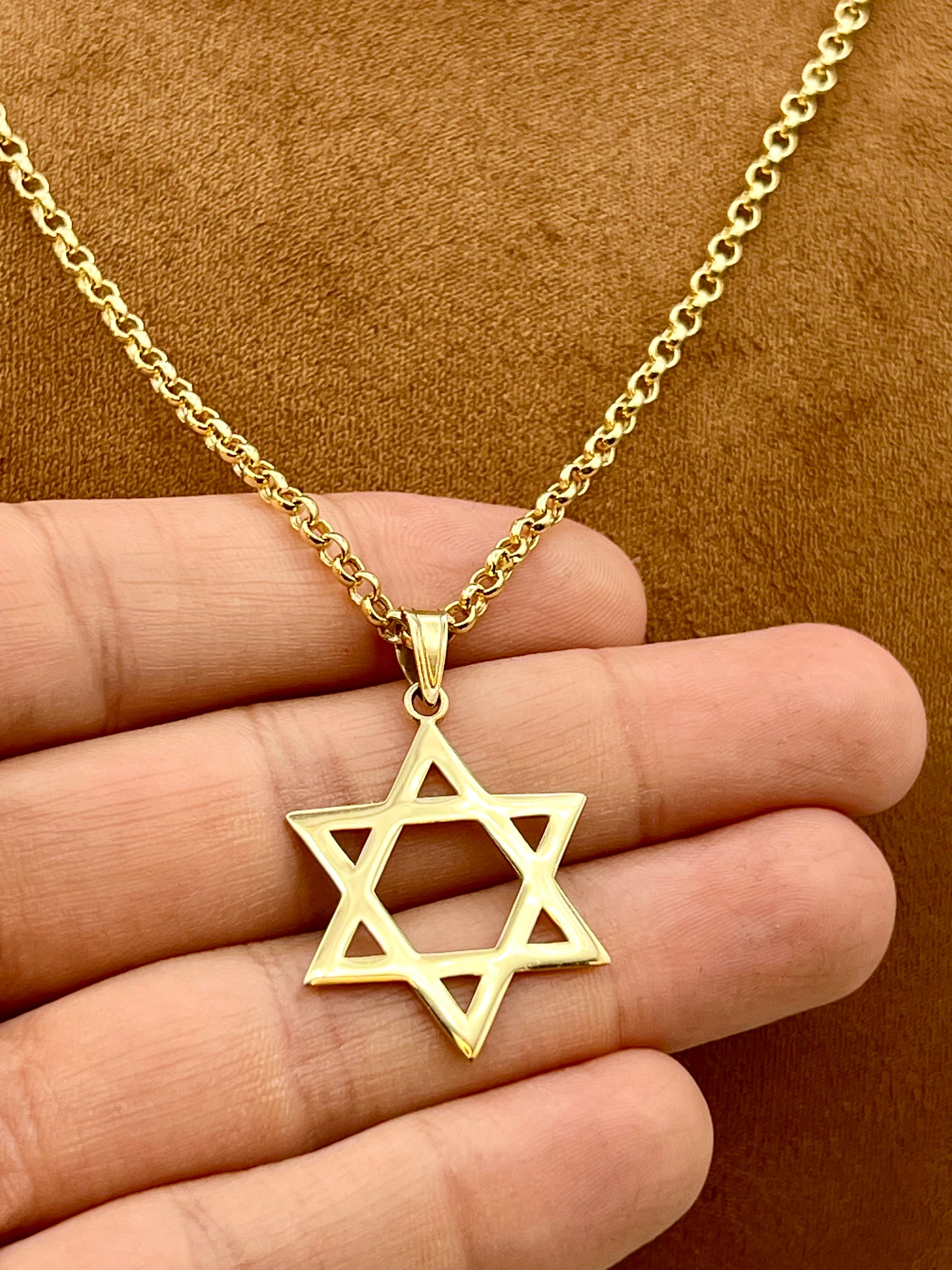 A gold Star of David pendant necklace held in a person's hand against a brown textured background.