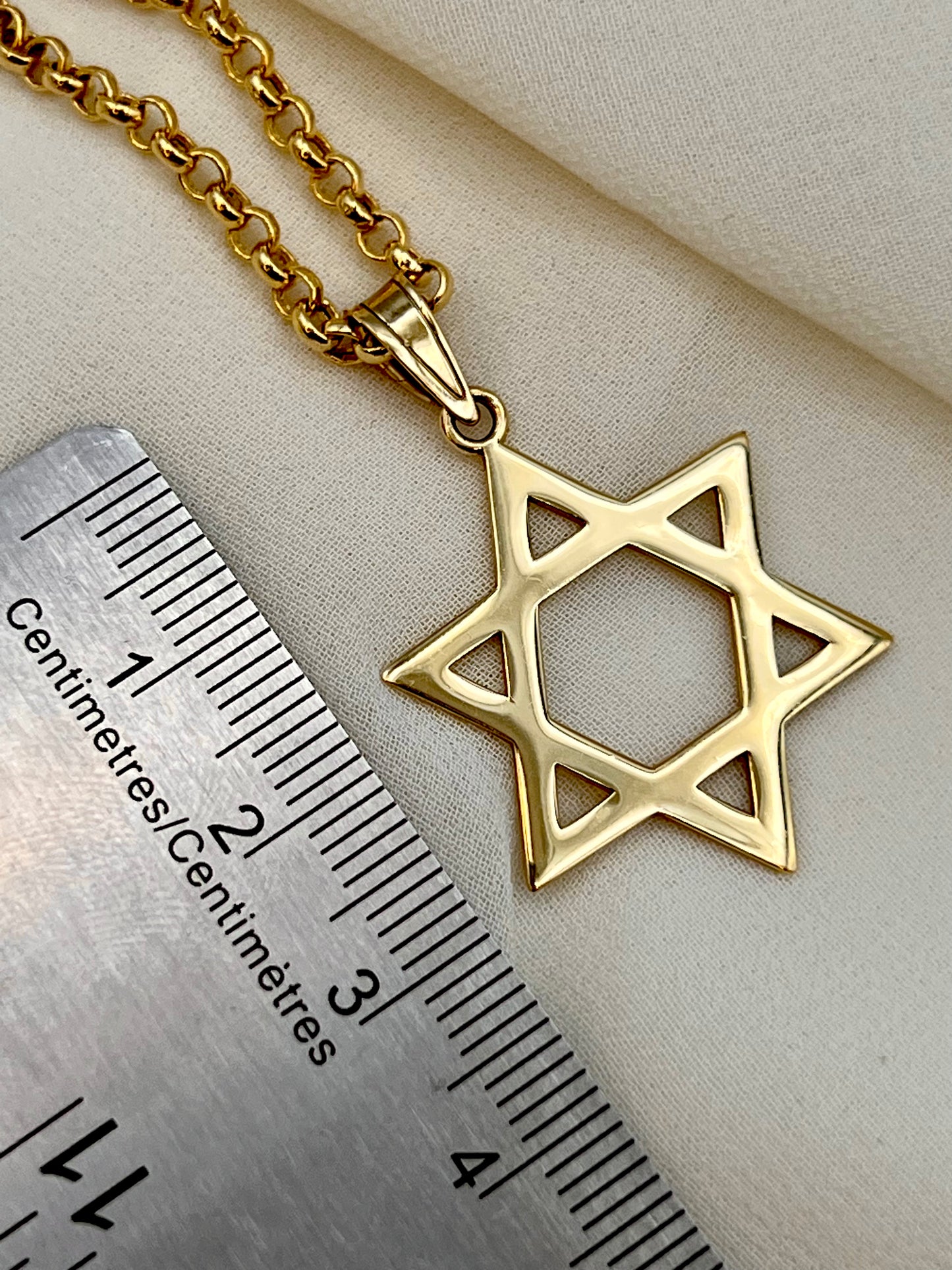 A gold Star of David pendant necklace on a matching chain, displayed next to a metal ruler to show scale on a beige fabric background.