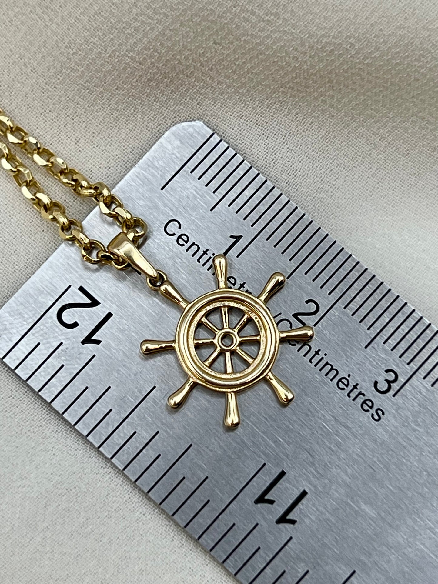 A gold ship's wheel pendant necklace on a matching chain, shown next to a metal ruler for scale on a beige fabric background.