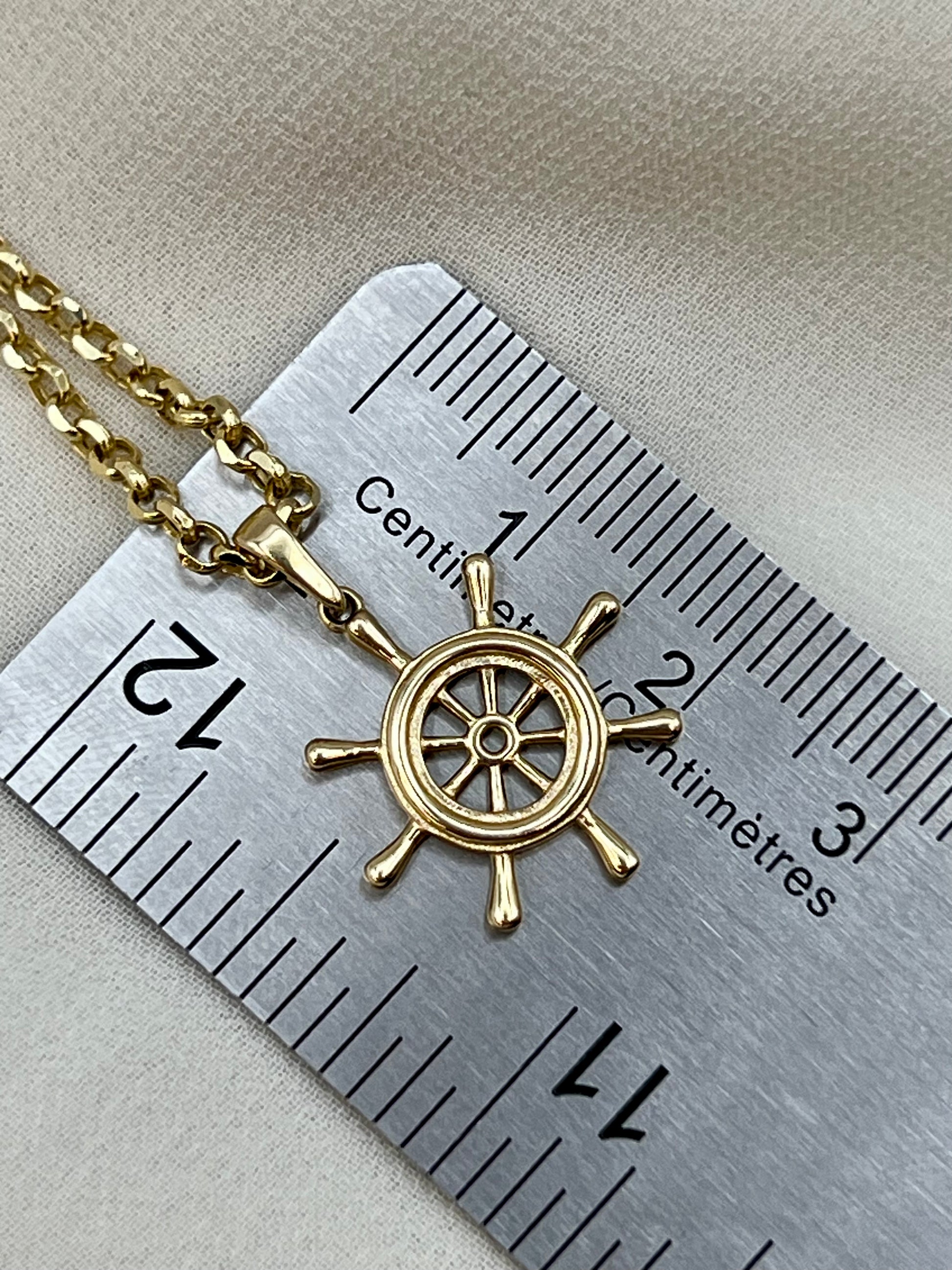 A gold ship's wheel pendant necklace on a matching chain, shown next to a metal ruler for scale on a beige fabric background.