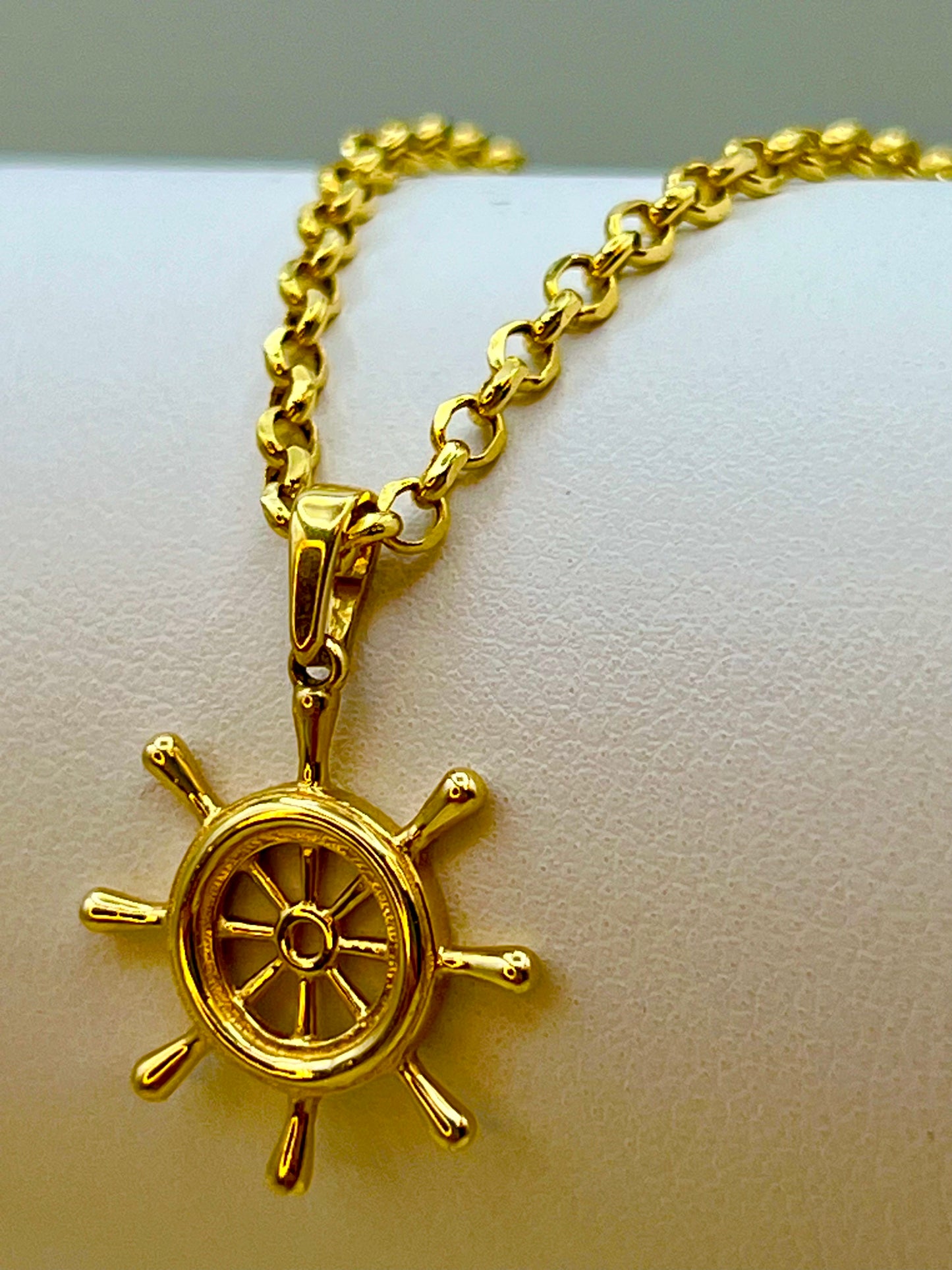 A gold ship's wheel pendant necklace displayed on a white textured surface, with the chain extending out of the top of the frame.