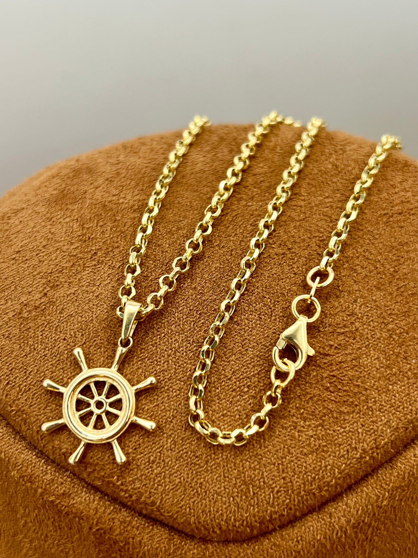 A gold necklace with a ship's wheel pendant, displayed on a brown textured velvet jewelry stand against a neutral gray background.