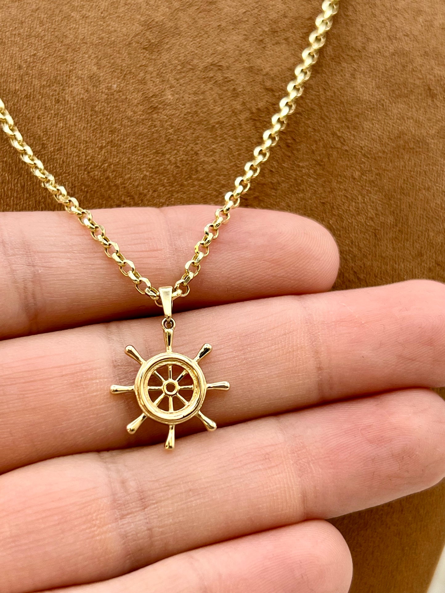 A gold ship's wheel pendant necklace held on a person's fingers against a brown textured background.